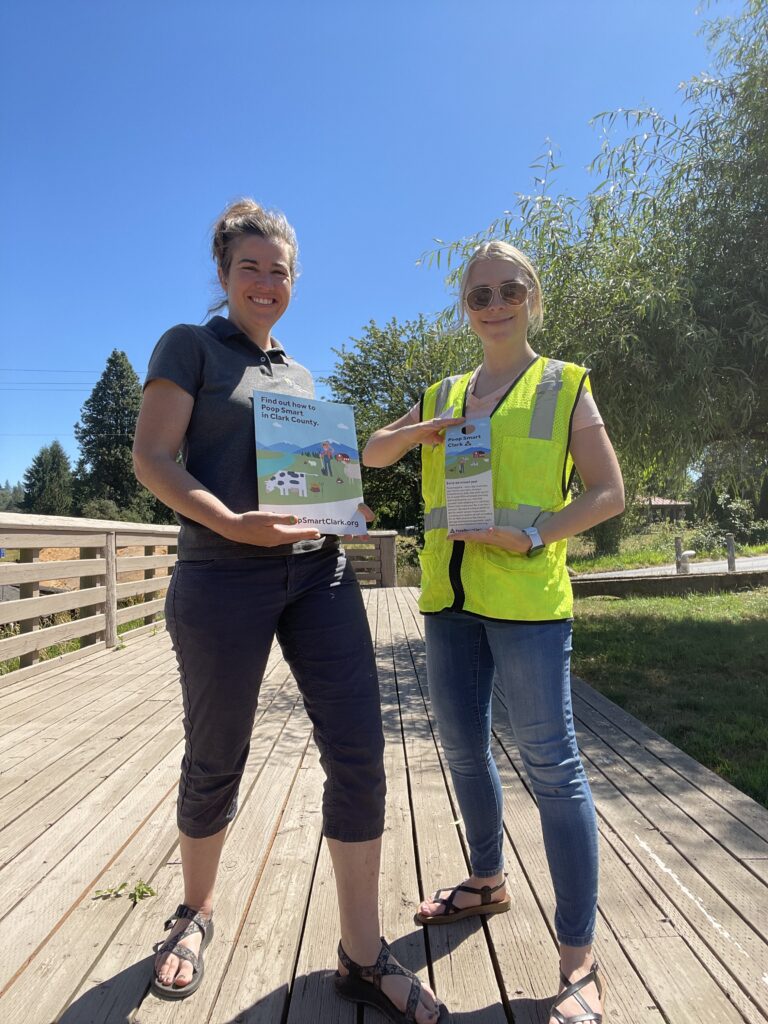 Two women stand on a boardwalk holding up paper materials for the Poop Smart Clark program. The woman on the left is holding one of the program’s informational packets and the woman on the right is holding one of the program’s door hangers.